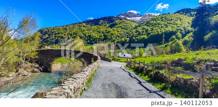 Scenic view of a winding road leading to a stone bridge over a river in the Pyrenees mountains with lush greenery and blue sky Scenic view of a winding road leading to a stone bridge over a river in the Pyrenees mountains with lush greenery and blue sky 140112053