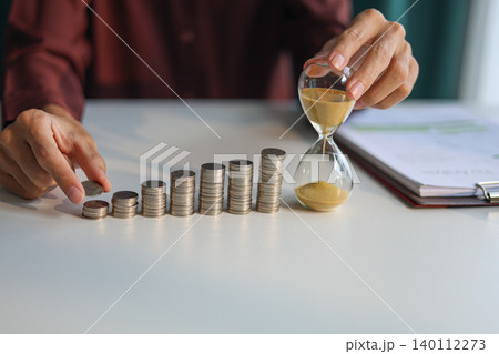 Time and money management concept. Rising stack of coins and running sand clock on the table. Growing of income, saving or return investment through time.  140112273