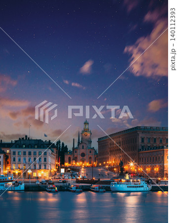 night view overlooking the illuminated historic architecture of Gamla Stan reflecting in the calm waters of Stockholm, Sweden, under a starry night sky 140112393