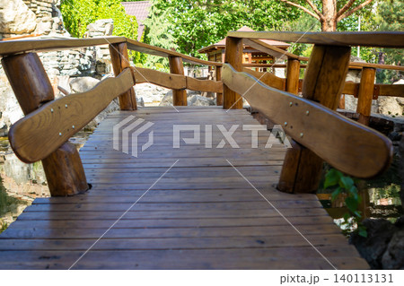 Wooden garden bridge over pond in a rustic park walkway surrounded by trees and stone features Wooden garden bridge over pond in a rustic park walkway surrounded by trees and stone features 140113131