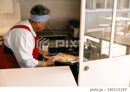 Senior Black woman chef baking fresh pizza in professional kitchen oven during busy day Senior Black woman chef baking fresh pizza in professional kitchen oven during busy day 140113197