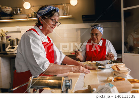 Active Caucasian and Black senior women rolling dough in a bright commercial kitchen setting 140113539
