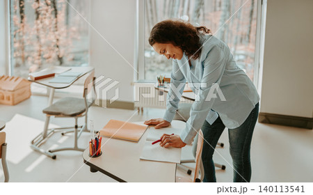 Mature Hispanic woman teacher grading student papers at desk in bright school classroom Mature Hispanic woman teacher grading student papers at desk in bright school classroom 140113541