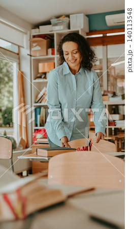 Smiling middle aged Hispanic female teacher arranging wooden chairs in a bright classroom Smiling middle aged Hispanic female teacher arranging wooden chairs in a bright classroom 140113548