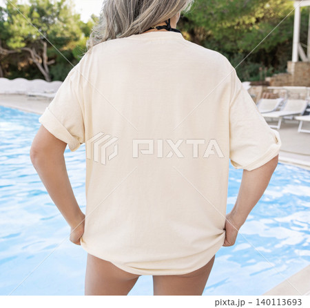 Blank light yellow oversized t-shirt mockup, woman poolside at sunset, back view with copy space 140113693