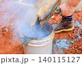 Someone pours dry mixture into bucket at construction site while smoke comes out dust is present on ground. 140115127
