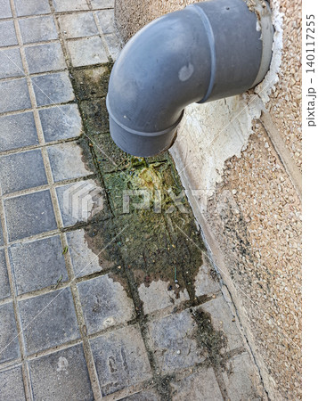 Grey plastic drainpipe leaks dirty water onto stone tiles. Green moss and algae grow alongside the damp area. This shows property maintenance issues in need of attention. Grey plastic drainpipe leaks dirty water onto stone tiles. Green moss and algae grow alongside the damp area. This shows property maintenance issues in need of attention. 140117255