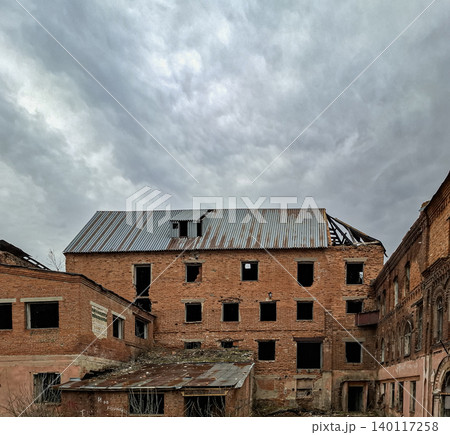 A vintage red brick industrial building stands empty and abandoned. Its metal roof is damaged, and stormy clouds fill the sky. Signs of decay cover the structure, hinting at past use. A vintage red brick industrial building stands empty and abandoned. Its metal roof is damaged, and stormy clouds fill the sky. Signs of decay cover the structure, hinting at past use. 140117258
