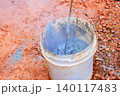 Worker stirs concrete mix in bucket at construction site with tools on dirt surface. 140117483