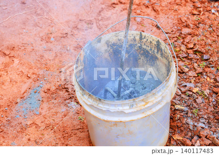 Worker stirs concrete mix in bucket at construction site with tools on dirt surface. Worker stirs concrete mix in bucket at construction site with tools on dirt surface. 140117483