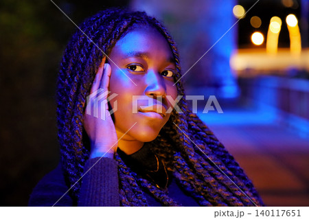 Moody night portrait of a young Black woman with long braids illuminated by vibrant blue and orange neon lights Moody night portrait of a young Black woman with long braids illuminated by vibrant blue and orange neon lights 140117651
