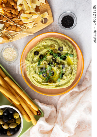 Broccoli hummus is artistically displayed in a bowl, garnished with olives. Accompanied by a charcuterie board featuring cheese, nuts, and breadsticks for a delightful snack experience Broccoli hummus is artistically displayed in a bowl, garnished with olives. Accompanied by a charcuterie board featuring cheese, nuts, and breadsticks for a delightful snack experience 140121286