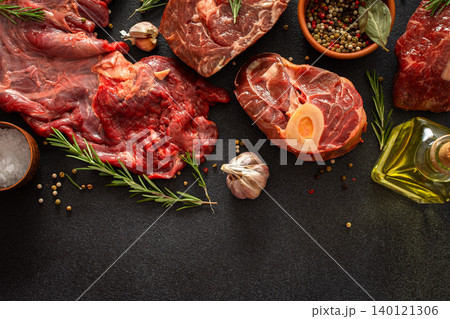 Different cuts of beef steak are arranged on a black surface. Fresh herbs, spices, and garlic accompany the meat, ready for cooking or grilling at a kitchen or market 140121306