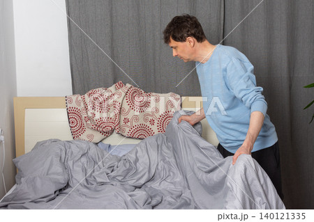 A 53-year-old man is actively changing the bed linen in his bedroom. He is focused on putting fresh sheets on the bed, demonstrating a common home chore A 53-year-old man is actively changing the bed linen in his bedroom. He is focused on putting fresh sheets on the bed, demonstrating a common home chore 140121335