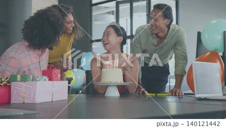 Celebrating team gathering around seated woman with tiara and red dress in office with cake, laptop Celebrating team gathering around seated woman with tiara and red dress in office with cake, laptop 140121442