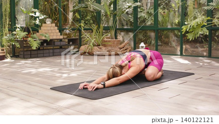 Stretching woman wearing pink sportswear folding forward on black mat in greenhouse with wristwatch Stretching woman wearing pink sportswear folding forward on black mat in greenhouse with wristwatch 140122121