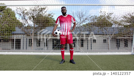 Standing adult soccer player holding black-and-white ball at hip before goal on turf, in red kit Standing adult soccer player holding black-and-white ball at hip before goal on turf, in red kit 140122357
