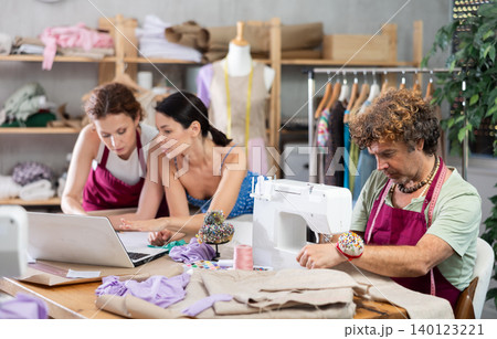 Two female dressmakers discussing dress designs on laptop. Man works on sewing machine 140123221