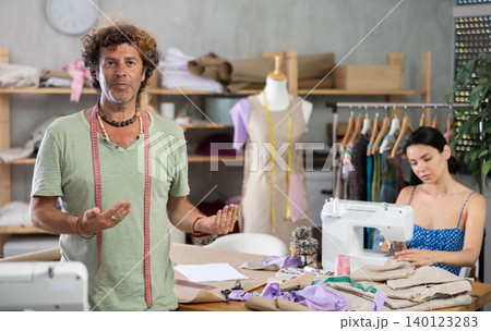 Middle-aged male dressmaker inviting clients while girl sew Middle-aged male dressmaker inviting clients while girl sew 140123283