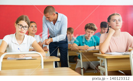Focused teenage students studying in classroom with teacher, writing lectures in workbooks Focused teenage students studying in classroom with teacher, writing lectures in workbooks 140123312