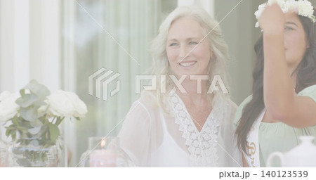 Smiling senior woman sitting in lace blouse at dining table, friend adjusting floral crown and sash 140123539