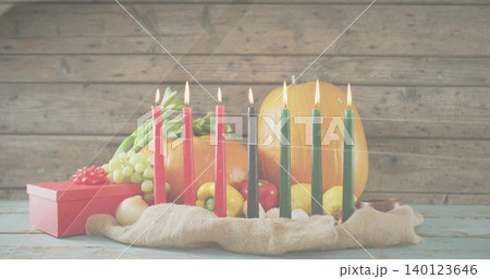 Displaying kinara seven lit candles glowing on worn pale-blue tabletop with pumpkin grapes red gift 140123646