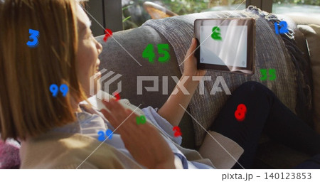 Waving Chinese woman holding tablet sitting on couch at home in beige sweater with numbers overlay 140123853