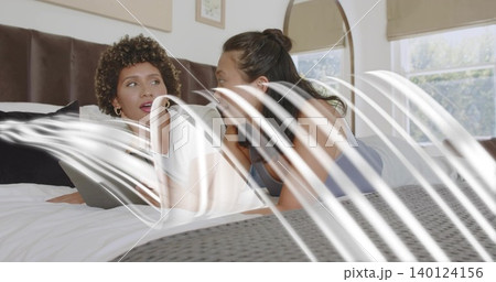 Chatting women wearing white top and gray tank reclining on bed, holding tablet by headboard Chatting women wearing white top and gray tank reclining on bed, holding tablet by headboard 140124156