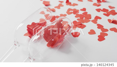 Tipping two clear stemmed glasses spilling many red heart confetti pieces across white tabletop 140124345