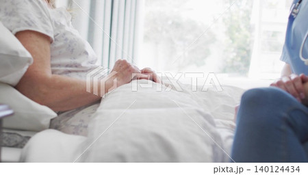 Lying patient resting hands on duvet at bedside, caregiver in blue scrubs with stethoscope watching 140124434