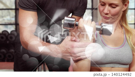 Guiding coach assisting woman in gray sports bra pressing chrome dumbbell in gym his hands spotting Guiding coach assisting woman in gray sports bra pressing chrome dumbbell in gym his hands spotting 140124492