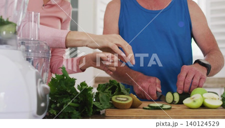 Slicing man blue tank at kitchen counter, reaching woman pink top, with cucumber and cutting board Slicing man blue tank at kitchen counter, reaching woman pink top, with cucumber and cutting board 140124529