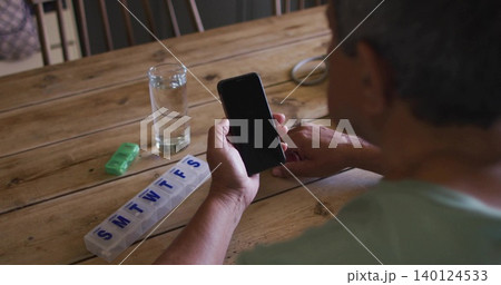 Holding smartphone, senior wearing teal shirt inspecting pill organizer at home table, copy space Holding smartphone, senior wearing teal shirt inspecting pill organizer at home table, copy space 140124533