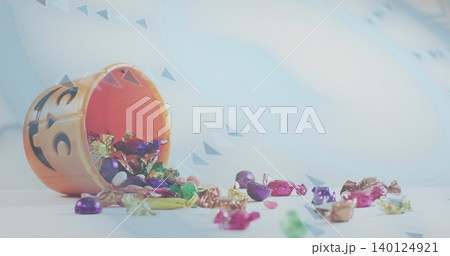 Tipping orange jack-o'-lantern bucket spilling foil candies across pale tabletop, confetti backdrop Tipping orange jack-o'-lantern bucket spilling foil candies across pale tabletop, confetti backdrop 140124921
