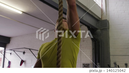 Climbing man wearing bright lime-green athletic T-shirt pulling upward at gym, with braided rope Climbing man wearing bright lime-green athletic T-shirt pulling upward at gym, with braided rope 140125042