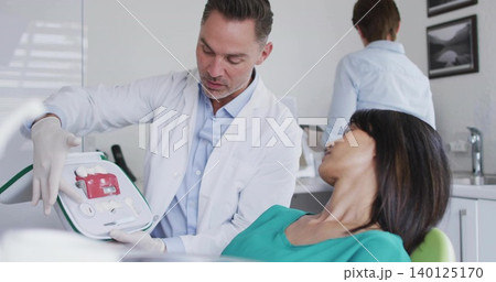 Kneeling practitioner in white coat, gloves holding portable AED with red pads in exam room 140125170