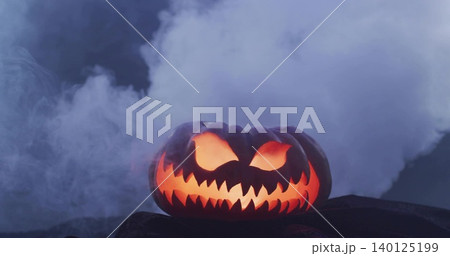 Glowing carved jack-o'-lantern casting warm orange light on dark rocky surface, with fog Glowing carved jack-o'-lantern casting warm orange light on dark rocky surface, with fog 140125199