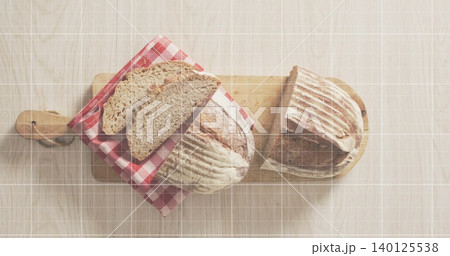 Showing two rustic sourdough loaves resting on light wood table, with cutting boards, gingham cloth 140125538