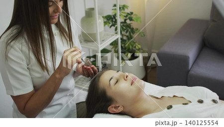 Applying practitioner wearing white uniform tinting client eyebrows on spa bed, with jar and brush Applying practitioner wearing white uniform tinting client eyebrows on spa bed, with jar and brush 140125554