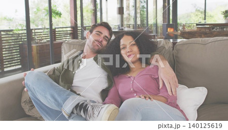 Sitting couple wearing casual clothes cuddling on light-beige sofa in home lounge, with glass doors 140125619