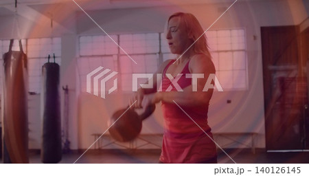 Swinging woman in red tank top and dark shorts lifting kettlebell in gym, with punching bags 140126145