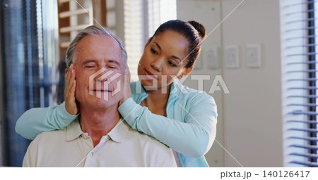 Standing caregiver massaging seated senior's neck near blinds, light-blue zip-up, pale polo shirt Standing caregiver massaging seated senior's neck near blinds, light-blue zip-up, pale polo shirt 140126417