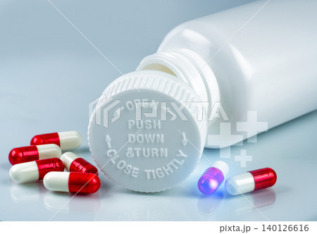 Red and white capsule pills next to plastic bottle with a childproof cap. Prescription medication, packaging safety, and healthcare control. Pharmaceutical innovation in modern packaging. Child safety Red and white capsule pills next to plastic bottle with a childproof cap. Prescription medication, packaging safety, and healthcare control. Pharmaceutical innovation in modern packaging. Child safety 140126616