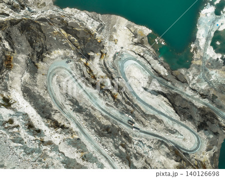 Aerial view of a transport truck hauling raw limestone through an open pit quarry, showing industrial mining operations and the environmental impact of material supply for global construction demand. Aerial view of a transport truck hauling raw limestone through an open pit quarry, showing industrial mining operations and the environmental impact of material supply for global construction demand. 140126698