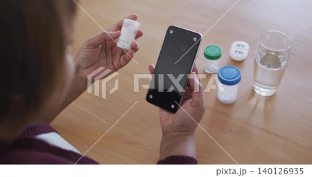 Mature woman in maroon sweater holding pill bottle and checking smartphone at home table with water 140126935