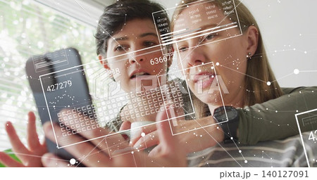 Examining two women leaning close and tapping smartphone by window, casual tops, smartwatch, AR UI Examining two women leaning close and tapping smartphone by window, casual tops, smartwatch, AR UI 140127091