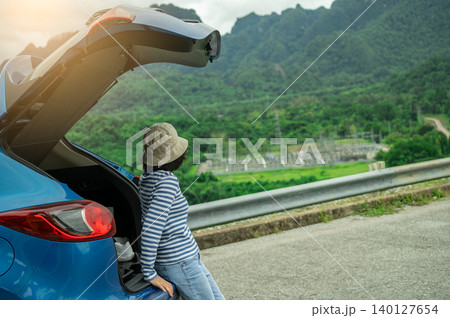 Asian woman enjoying peaceful mountain vista during sustainable road trip adventure with open SUV car tailgate on eco friendly summer vacation exploring green forest landscape for wellness retreat. Asian woman enjoying peaceful mountain vista during sustainable road trip adventure with open SUV car tailgate on eco friendly summer vacation exploring green forest landscape for wellness retreat. 140127654