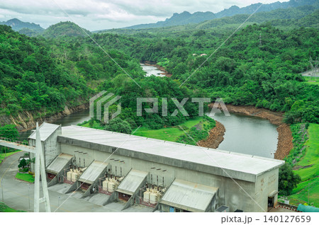 Hydropower plant building with green forest and high-voltage grid infrastructure, with mountain landscape in background. Hydroelectric power station at dam site. Renewable energy. Power generation. 140127659