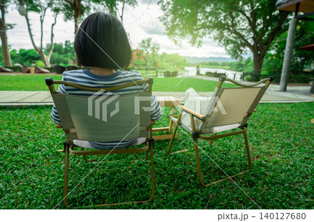 Rear view of a woman relaxing on lawn chair by river. Slow living and personal retreat. Digital detox weekend with nature and greenery. Mental wellness, healing, and nature-connected lifestyle. Rear view of a woman relaxing on lawn chair by river. Slow living and personal retreat. Digital detox weekend with nature and greenery. Mental wellness, healing, and nature-connected lifestyle. 140127680