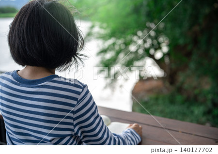 Rearview of a woman sitting on a balcony reading a book, representing a digital detox retreat, relaxation, and mindfulness practice for mental wellness, self care, and peaceful lifestyle balance. 140127702